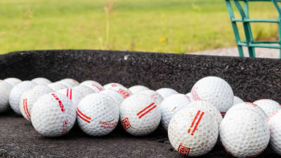 Flera golfbollar bredvid varandra på en grå yta. I bakgrunden syns gräs och en grön korg.