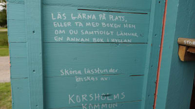 Ett turkost plank med vit text där det står: "Läs gärna på plats eller ta med boken hem om du samtidigt lämnar en annan bok i hyllan.