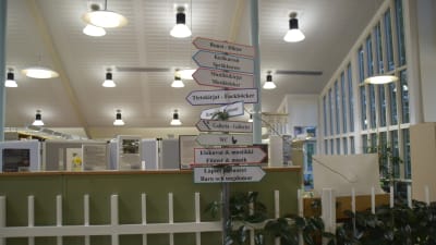 Hemgjorda textade skyltar i ett bibliotek som pekar åt vilket håll bland annat barnböcker, fackböcker och filmer finns. Många skyltar på en stolpe, mitt i ett bibliotek.