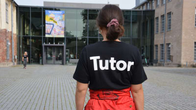 En kvinna med röd halare och en svart t-skjorta med texten tutor på ryggen står på Arkens gård.