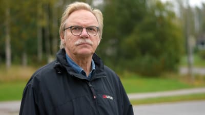 En äldre man med mustasch och glasögon. Står ute. Klädd i mörkblå jacka.