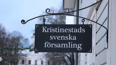 En skylt hänger från väggen. På skylten står det Kristinestads svenska församling.