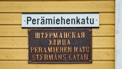 Keltaisessa puutalossa katukyltti jossa teksti Perämiehenkatu, alla kyltti jossa venäjänkielistä tekstiä