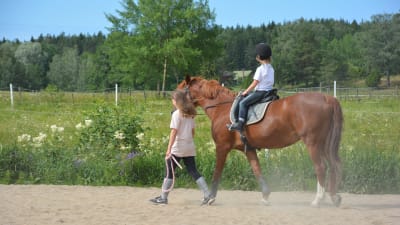 En tolvårig flicka leder en häst som en mindre flicka rider på, på en sandplan.