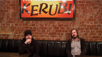 Musikerna Aki Tykki och Ville Karttunen sitter i svart lädersoffa, ovanför dem neonskylt med texten Ravintola Kerubi.