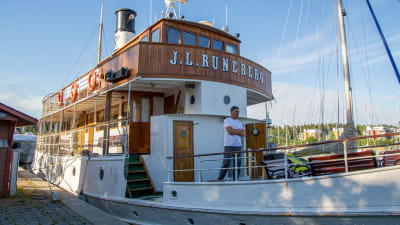 Båt som det står "J. L Runeberg" på förtöjd vid en hamn. En person står på däcket. 