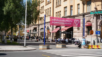 Ett stort övergångsställe framför en skylt där det står sommargågata. På gågatan sitter människor på uteserveringar.
