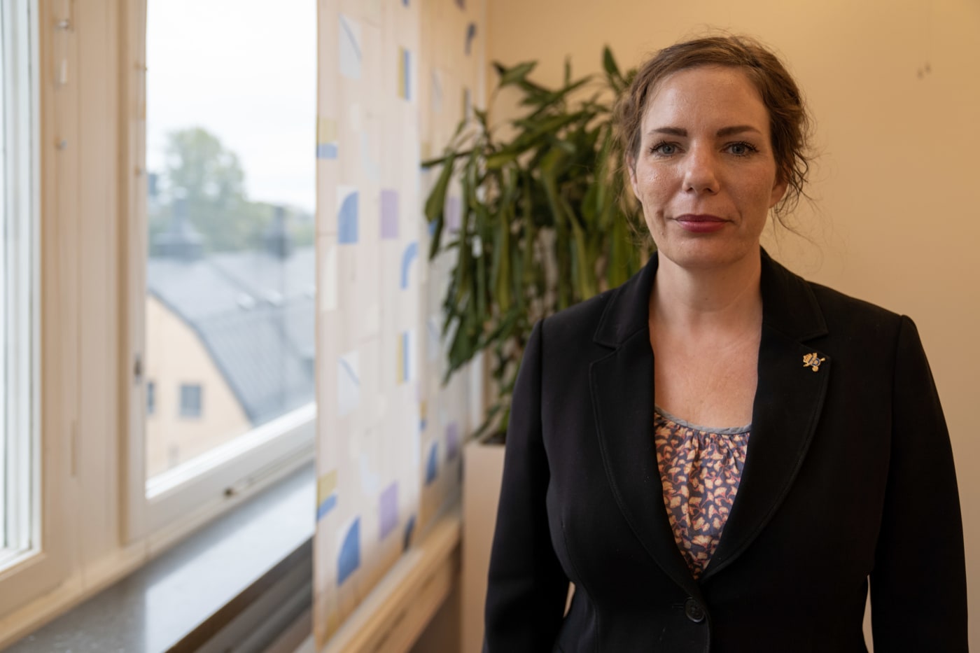 Sarah Pearson guarda nella telecamera. Lei è in piedi vicino alla finestra con una giacca.