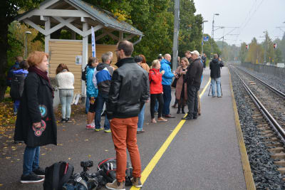 Människor står på en tågperrong, tågbana till höger, höstlöv på marken.