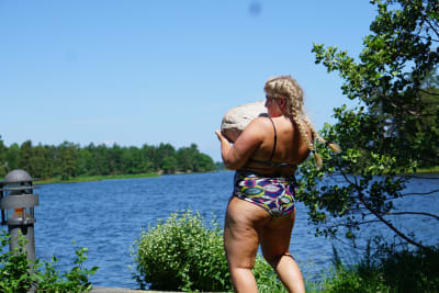 Sara Dahlström iklädd bikini. Det är sommar och hon står vid en sjö eller hav. Hon lyfter en stor sten. 