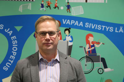 Man i kostymjacka står framför mural som avbildar utbildning och fostran