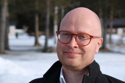En man med runda röda glasögon. I bakgrunden syns träd och snö.
