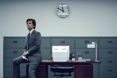 En allvarlig Mark S. (Adam Scott) med ett plåster på pannan, sitter på kanten av ett skrivbord i ett grått rum iklädd en grå kostym.