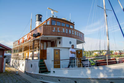 Båt som det står "J. L Runeberg" på förtöjd vid en hamn. En person står på däcket. 