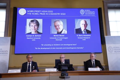 Tre män sitter vid ett skrivbord. I bakgrunden en powerpoint som visar vinnarna av Nobelpriset i kemi.
