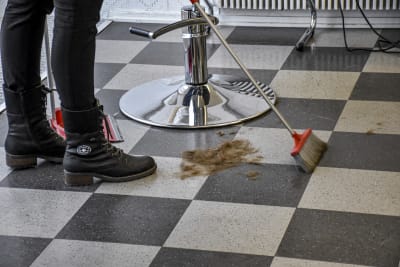 En person med svarta skor sopar upp hårstrån som ligger nedanför en frisörstol.