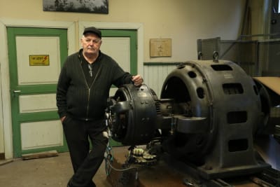 En man med keps på huvudet står och lutar sig mot en gammal generator.