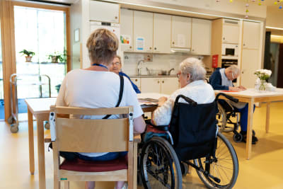 Två kvinnor vid ett bord, med ryggen mot kameran, den ena med grått hår, sittande i rullstol. 