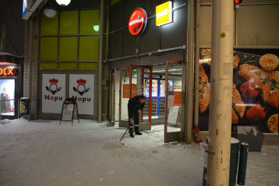En man i väktarkläder sopar bort snön framför dörrarna till en stor affär.