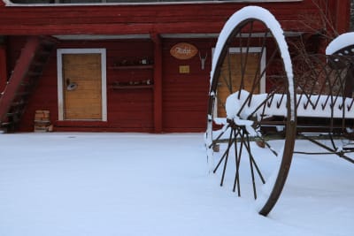 I förgrunden en gammal snöbeklädd harv. I bakgrunden en röd träbyggnad.