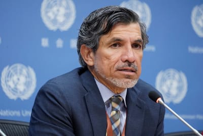 En man med grått hår och kort skägg talar i en mikrofon. På väggen bakom honom upprepas FN:s logotyp.