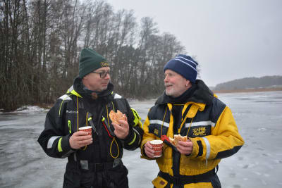 Två män står på isen, dricker kaffe ur juliga pappmuggar och äter wienerbröd. De har flytdräkter, en svart och en gul overall på sig, och isdubbar runt halsen.