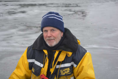 En man med gråvitt skägg och mustasch, blå mössa, gulsvart overall och isdubbar runt halsen står på isen och tittar in i kameran.