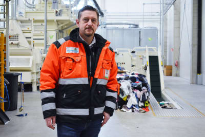 En brunhårig man i orange-svart rock står inne i en fabriksbyggnad.