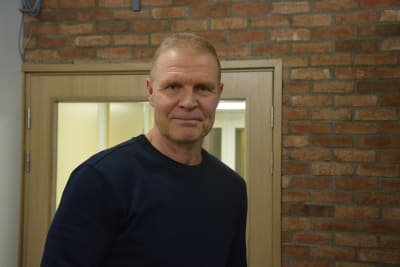 Man i 50-60-årsåldern står framför en dörr och en tegelvägg och tittar in i kameran.