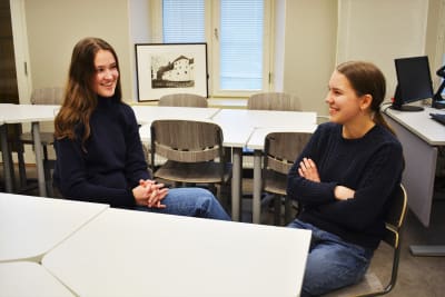 Två flickor i tonårsåldern sitter och pratar i ett klassrum.
