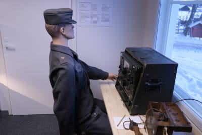 En mannekäng har placerats sittande vid ett bord framför en radioapparat. Dockan har militära kläder från 1940-talet på sig.