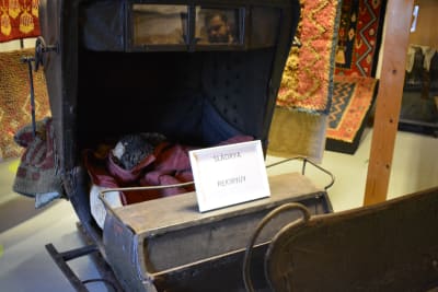 En gammal, svart, täckt släde på ett museum, med en vinröd-svart rya som täcke, en så kallad slädrya.