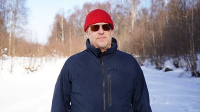 En man med blå jacka, röd mössa och solglasögon står på isen och tittar in i kameran.