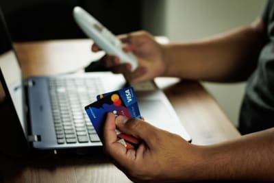 En person sitter vid en bärbar dator och håller i två kreditkort i ena handen och en telefon i andra handen.