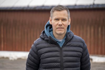 En man med gråsprängt kort hår och mörkblå tunn dunjacka står ute. I bakgrunden en byggnad med rödmålad träfasad.
