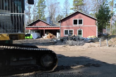 Grävmaskin i förgrunden och en stor röd träbyggnad i rivningsskede i bakgrunden.