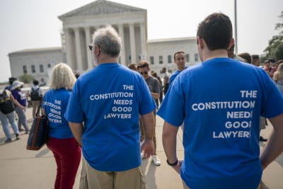 Människor väntar på beslut utanför Högsta domstolens byggnad i Washington. Tre personer bär blåa t-tröjor med trycket "The Constitution needs good lawers" (Konstitutionen behöver duktiga jurister) på ryggen.