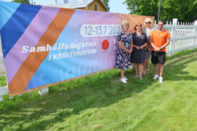 Tre kvinnor och en man poserar bredvid en stor banderoll. På banderollen finns texten Samhällsdagarna i Kristinestad 12-13.7.2023.
