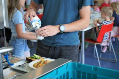 En ung kille häller upp mjök i ett glas. I bakgrunden elever som äter skollunch.