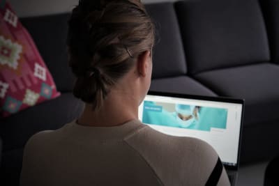 En kvinna med håret uppsatt i en knut sitter med ryggen mot kameran och en dator i famnen. På skärmen syns en läkare med munskydd.