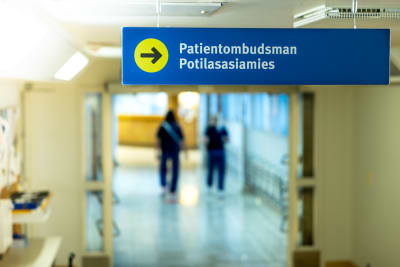 Skylt till patientombudsmannen, två blåklädda sköterskor går i bakgrunden