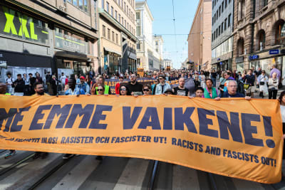 Människor i ett demonstration som tar upp hela gatans bredd, längst fram håller flera demonstranter upp ett stort tygstycke med texten "Vi tiger inte!" på finska.