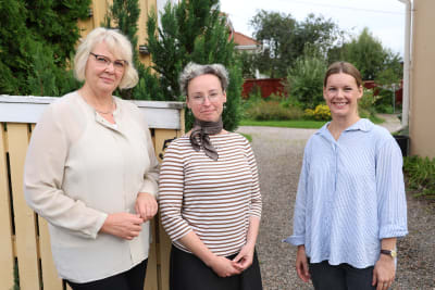Tre kvinnor står vid ett gult staket. I bakgrunden syns en lummig trädgård.