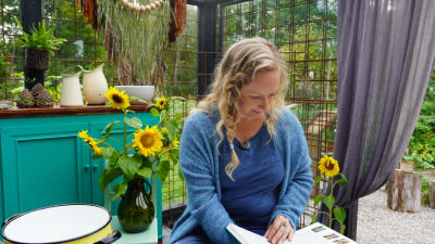 Alexandra De Paoli sitter i trädgårdshuset och läser en bok. Solrosor i en vas bakom henne.
