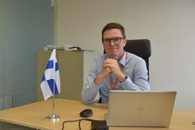 Bild på Lauri Raunio som är chef för Finlands handelscenter i Taiwan. Han sitter vid ett skrivbord med en dator och en finsk flagga.