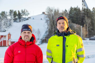 Två män som tittar in i kameran. I bakgrunden en skidbacke.