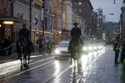 Sorgetåget på Alexandersgatan. Längst fram två rytare, därefter följer en lång rad bilar och ljuset från deras lyktor blänker på de regnvåta gatstenarna. 