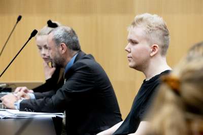 Aleksanteri Kivimäki fotograferad från sidan medan han sitter vid ett svart bord i en rättssal. Bredvid honom sitter en man och en kvinna.