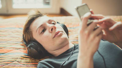 En kvinna med kort hår ligger på en säng och lyssnar på musik. Hon har hörlurar på sig och tittar på sin smarttelefon. 