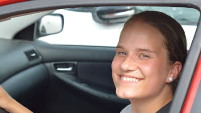 Ung tjej sitter i en bil och ler mot kameran.